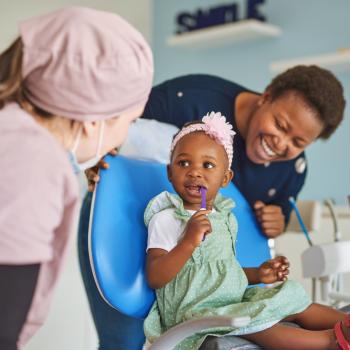 Uma menina está sentada na cadeira de uma dentista, com a escova de dentes na boquinha, observada pela sua mãe e a profissional. O cenário nos remete a importância da saúde bucal infantil