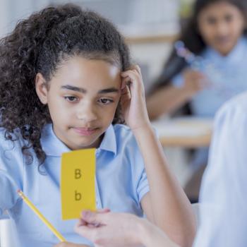 Imagem de uma criança com dislexia olhando para uma ficha amarela com letras B maiúscula e minúscula. Em uma mão ela segura um lápis e a outra está sobre a cabeça em uma postura de dúvida. 