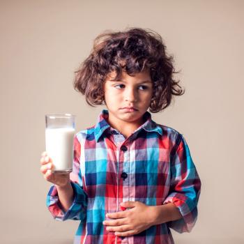 Imagem de uma criança desconfortável, com um copo de leite em uma mão e outra sobre a barriga em uma postura que remete aos efeitos da intolerância à lactose.