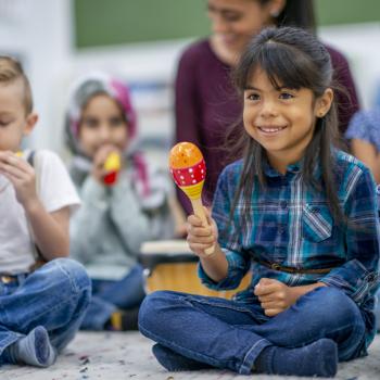 Em uma sala de aula temos algumas crianças participando de uma atividade com músicas. Em destaque, uma menina sentada no chão segurando um instrumento musical que lembra um chocalho. A imagem remete ao tema musicalização