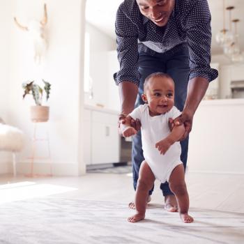 Um pequeno bebê é incentivado pelo seu pai a caminhar pela sala de casa. O adulto acompanha a criança nessa atividade, segurando os seus bracinhos. Essa imagem remete ao estímulo dos primeiros passos do bebê