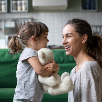 mãe e filha se olhando e sorrindo num ambiente familiar em frente ao sofá da casa, onde a criança abraça um ursinho de pelúcia e olha sorrindo para a mãe