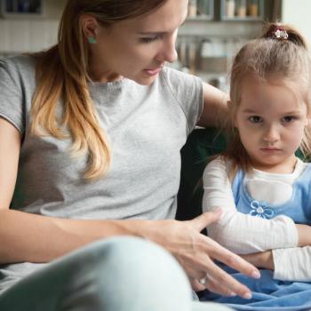 mãe e filha sentadas no sofá da sala, mãe abraça a criança e conversa, criança parece estressada com os braços cruzados e olhar distante