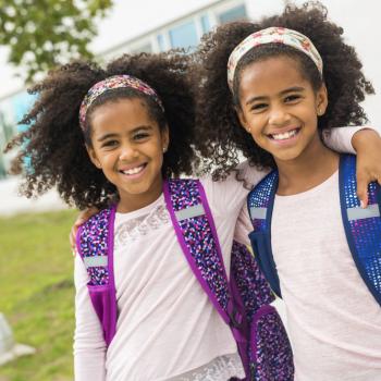 Duas irmãs gêmeas se abraçando lado a lado, sorridentes, com mochila nas costas, ao ar livre em frente a escola.
