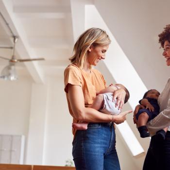 Duas mulheres adultas estão carregando seus respectivos bebês no colo, enquanto conversam, em um ambiente interno de uma residência