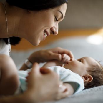 Imagem de uma mãe acariciando um bebê deitado. O carinho e a troca de olhares entre eles ajuda a fortalecer o vínculo entre os dois
