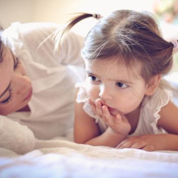 Mãe e filha deitadas na cama, onde a mãe alisa os cabelos da criança e a mesma aparenta estar reflexiva