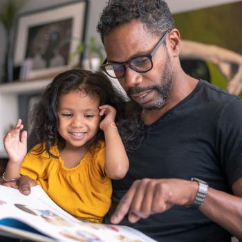 pai e filhas sentados juntos no sofá da sala lendo um livro 