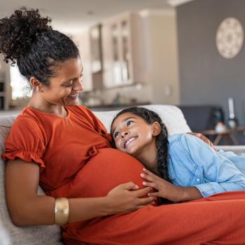 Mãe grávida e filha abraçada em sua barriga, ambas se olham e sorriem, sentadas no sofá da sala de casa.