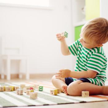 Criança sentada brincando sozinha com dadinhos no tapete da salinha de brinquedos.