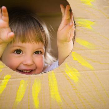criança sorrindo dentro de uma caixa de papelão, seu rostinho está na parte interior da caixa, num recorte circular onde foi desenhado um sol