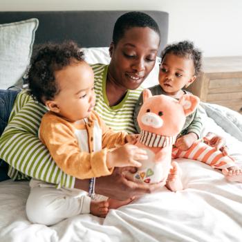 Mãe com dois filhos no colo, sentados em cima da cama enquanto brincam com um ursinho de pelúcia