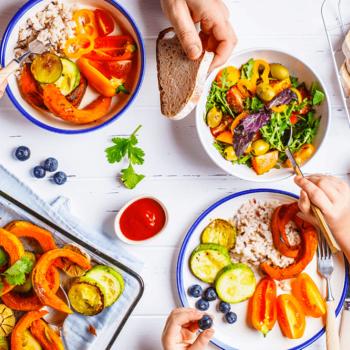 Foto tirada de cima de uma mesa com a alimentação familiar. Há mãos de adulto e de criança e elas se servem de diversos alimentos alis dispostos: pães, frutas, cereais e legumes.