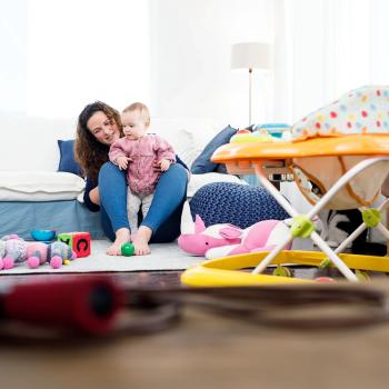 Ao fundo temos uma mãe brincando com um bebê e o estimulando a ficar em pé, ao lado temos brinquedos e um andador infantil