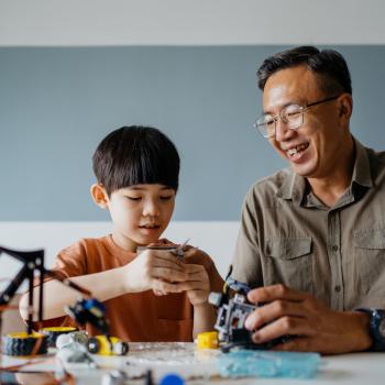 Um homem com traços asiáticos junto de seu filho com peças robóticas nas mãos montando algo.