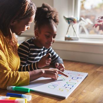 Mulher negra cabelo castanho escuro e vestindo uma blusa amarela ao lado de sua filha negra de cabelo castanho escuro com uma blusa preta com listas brancas na horizontal, a mãe está ajudando a filha a ler um papel que está escrito as letras do alfabeto