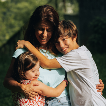 Uma mãe branca vestindo uma blusa azul esverdeado abraçando seus filhos, do lado direito um menino branco e loiro vestindo uma blusa branca e do lado esquerdo uma menina com síndrome de down utilizando um vestido branco com detalhes vermelhos