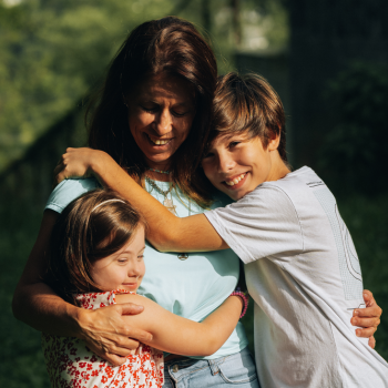 Aprender no Ninho: Gisele, Sara e tudo aquilo que as crianças podem nos ensinar Uma mãe branca vestindo uma blusa azul esverdeado abraçando seus filhos, do lado direito um menino branco e loiro vestindo uma blusa branca e do lado esquerdo uma menina com síndrome de down utilizando um vestido branco com detalhes vermelhos