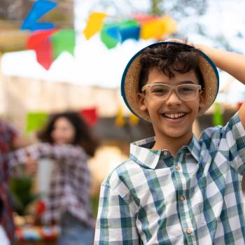 Foto de uma criança sorrindo, usando óculos de grau, camisa listrada e segurando um chapéu de palha. Ao fundo, festa junina, com bandeiras e adultos comemorando.