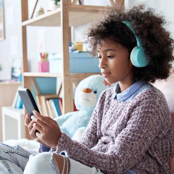 #ParaTodosVerem Menina negra pré-adolescente, com cabelo cacheado, solto, usando fones de ouvido azuis enquanto olha a tela do celular. Ela está sentada em uma cama com almofadas e veste blusão com calça jeans. O fundo da foto é um quarto de menina.