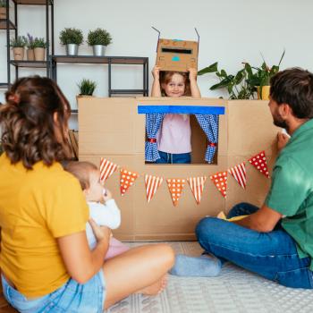 Um homem, uma mulher e um bebê estão sentados no chão de uma sala de estar, de costas, assistindo a uma apresentação de uma menina. Ela está dentro de uma caixa de papelão, simulando uma casinha.