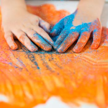 Imagem das mãos de uma criança, sentindo a textura de um glitter e de tintas. Ao fundo, um cartaz branco com algumas pinturas na cor laranja, rosa e glitter azul.