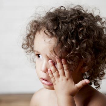 Uma criança, com cabelos cacheados de cor castanha com a mão no rosto, coçando o olho.