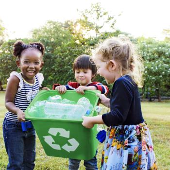 Imagem de três crianças, da esquerda para direita, uma menina, um menino e mais uma menina segurando um lixo com garrafas para reciclagem. Ao fundo, árvores e gramado.