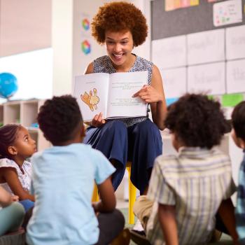 Descrição imagem: Foto de uma mulher segurando um livro e apresentando para as crianças em uma sala de aula sobre educação bilíngue.