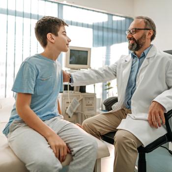 Fimose infantil: entendendo o “pintinho” dos meninos Imagem de um ambiente hospitalar em que um médico tranquiliza um paciente menino, ao colocar a mão no ombro dele. O médico está de jaleco branco, óculos e tem barba e o menino está vestindo uma camiseta azul