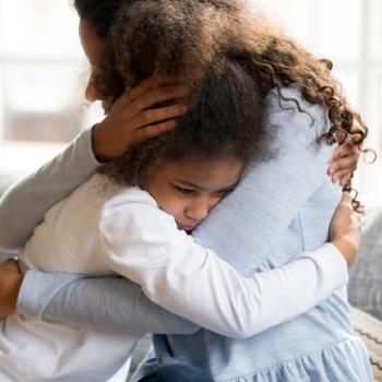 Sentadas em um sofá cinza, uma mulher, usando uma blusa azul clara, abraça uma menina, que usa uma blusa branca. A menina está triste.