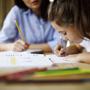 Sentados à mesa, estão uma mulher, que veste uma camisa azul, e uma menina, que veste uma camisa branca. A mulher e a menina estão escrevendo em cadernos, e a menina aparenta estar concentrada. Na mesa, estão alguns livros e canetas coloridas. 