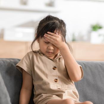 Dentro de uma casa, está uma menina sentada em um sofá cinza, usando um vestido marrom. A menina está com expressão de tristeza, com os olhos fechados e a mão apoiada na cabeça.