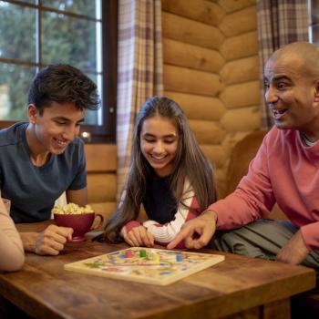 Dentro de uma casa, todos sentados em volta de uma mesa de madeira, está uma família composta por um homem, uma mulher, duas meninas e um menino. Com sorrisos e brincadeiras, eles estão jogando um jogo de tabuleiro.
