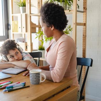 Parentalidade atípica com foco em neurodiversidade - Conheça o projeto Sentados à mesa para estudar, uma mulher, usando uma blusa rosa, conversa com um menino, que está usando fones de ouvido e uma camisa estampada, e parece estar entediado enquanto está debruçado em seu caderno. Na mesa estão um notebook, lápis, canetas coloridas e uma xícara.