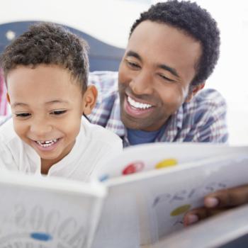 Deitados em uma cama, um homem, vestido com uma camisa quadriculada azul, está lendo um livro com um menino, que veste uma camisa branca. Com o livro aberto, o menino e o homem estão sorrindo. 