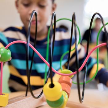 Em uma sala, com pessoas ao fundo, um menino, que está vestindo uma camisa com listras azuis, brancas, amarelas e marrons, está sentado à mesa, brincando com um brinquedo colorido, com formas geométricas.