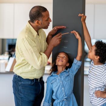 Marcos do desenvolvimento infantil: etapas cruciais do crescimento Em uma cozinha, está um homem, uma menina e um menino. A menina está encostada em uma parede, enquanto o homem e o menino marcam na parede qual é a altura dela.