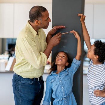 Em uma cozinha, está um homem, uma menina e um menino. A menina está encostada em uma parede, enquanto o homem e o menino marcam na parede qual é a altura dela.