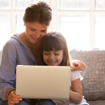 Sentadas em um sofá cinza, estão uma mulher e uma menina. A mulher está segurando um tablet. Sorridentes, ela e a menina assistem a um vídeo no dispositivo.