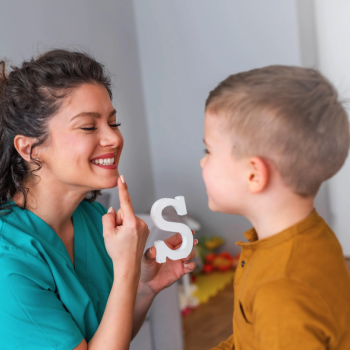 Uma mulher sorrindo, apontando para a própria boca e segurando um objeto em forma de letra “S”, enquanto uma criança observa e tenta imitar a ação da mulher.