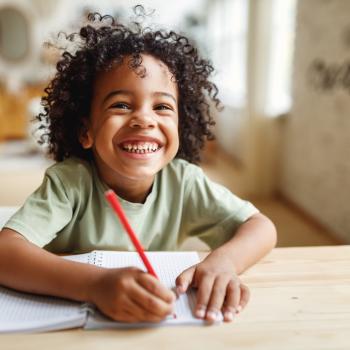 Descrição: criança sorrindo, segurando um lápis e um caderno para escrever. ao fundo, uma sala como ambiente.
