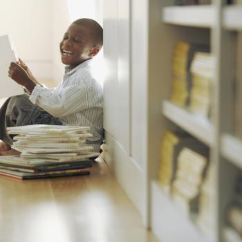 Um garoto está sentado no chão, ao lado de uma estante de livros. Ele sorri, lendo um livro. Ao seu lado, está uma pilha de outros livros.