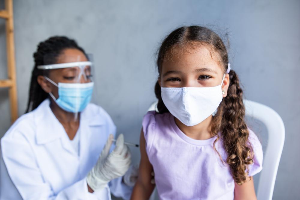 Um enfermeira de máscara e proteção facial realiza a vacinação infantil. Ela aplica a vacina com uma seringa em uma garotinha que está sentada e também usa máscara.  