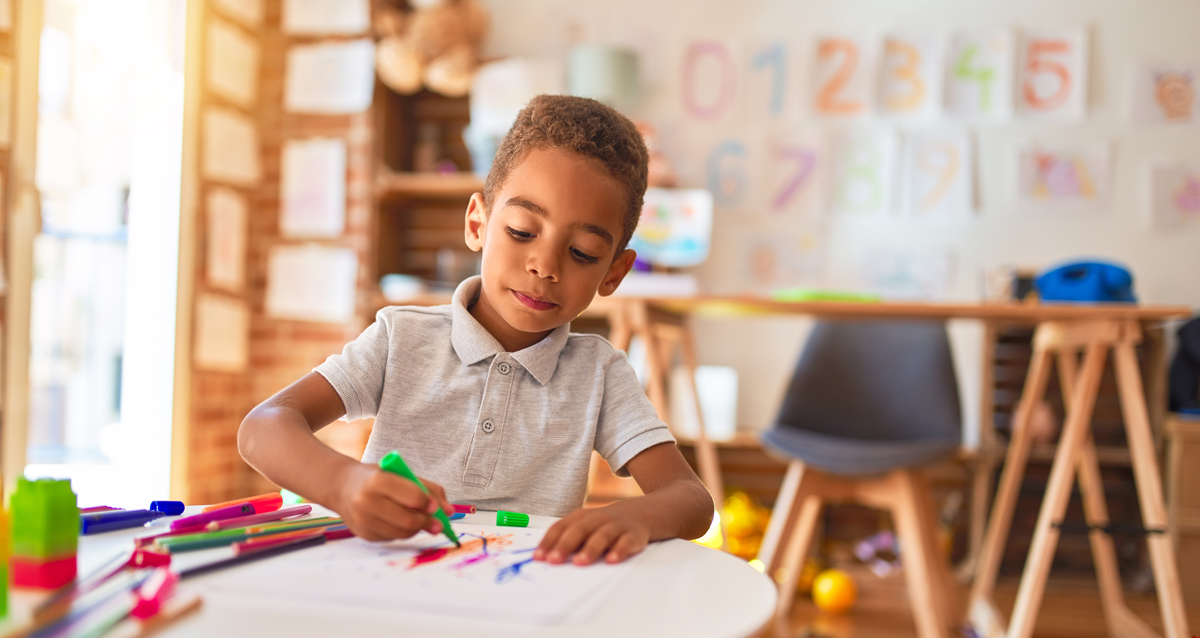 Uma criança de 4 anos está em uma sala de aula, sentado à mesa, e usa uma caneta hidrocor para desenhar. 