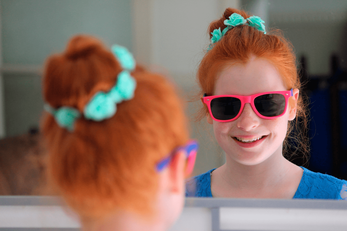 Menina sorri para a própria imagem no espelho. Se trata de uma criança de cerca de 9 anos usando óculos escuros com armação rosa.
