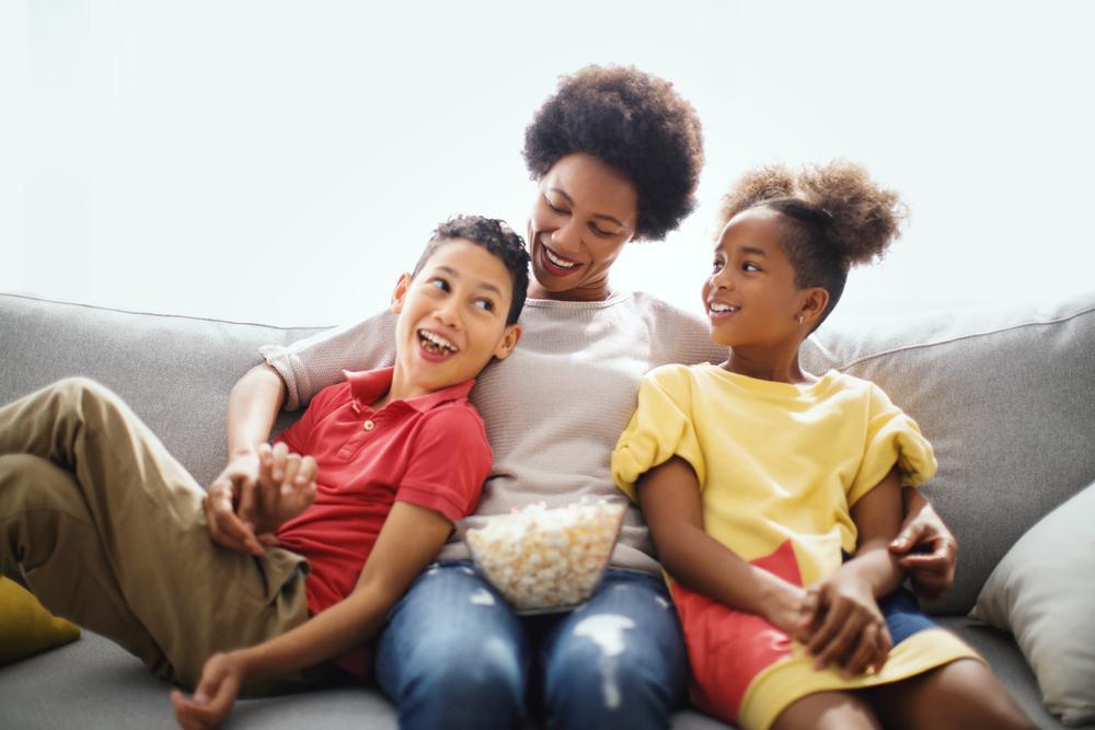 Mãe, com uma vasilha de pipoca no colo, abraça filho e filha sentados em um sofá para assistir filmes sobre maternidade. Todos sorriem.