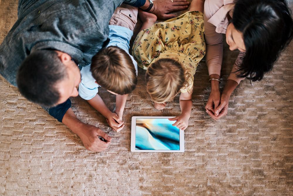 Tempo de tela: pai, mãe e duas crianças pequenas estão deitados em um tapete e olham juntos para a tela de um tablet.