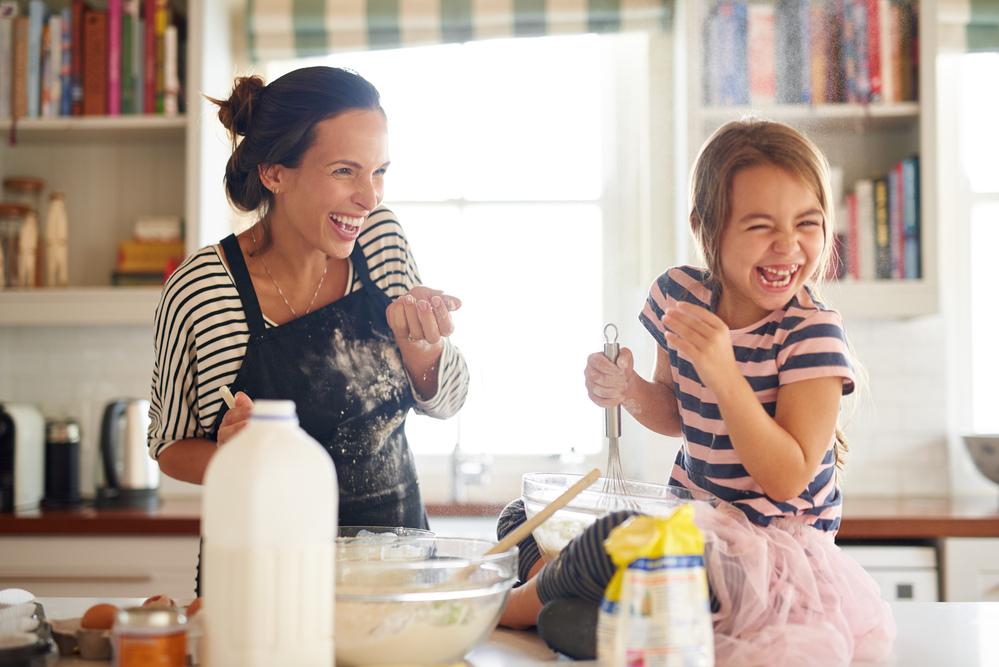 Mãe e filha cozinham juntas em uma bancada de cozinha e dão risada, ilustrando um dos hábitos que favorecem uma alimentação saudável. 