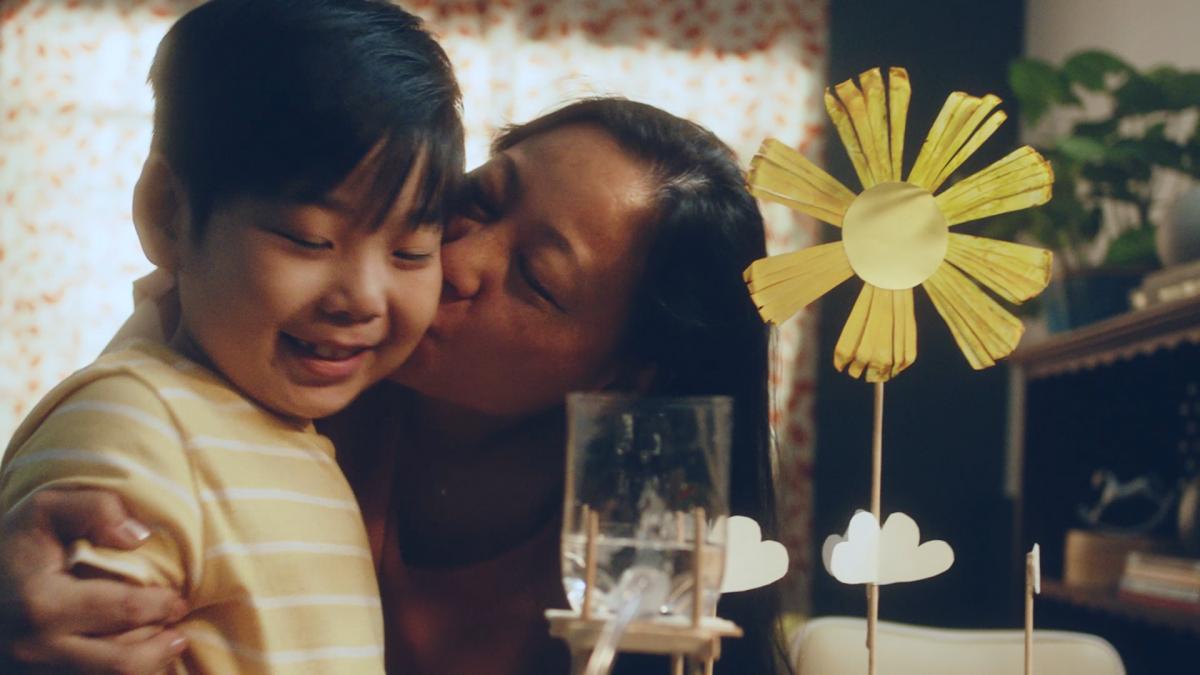 Representando a relação de mães e filhos, a foto mostra uma mãe beijando seu filho na bochecha enquanto ambos sorriem. Em frente deles, há uma flor feita de papel. 